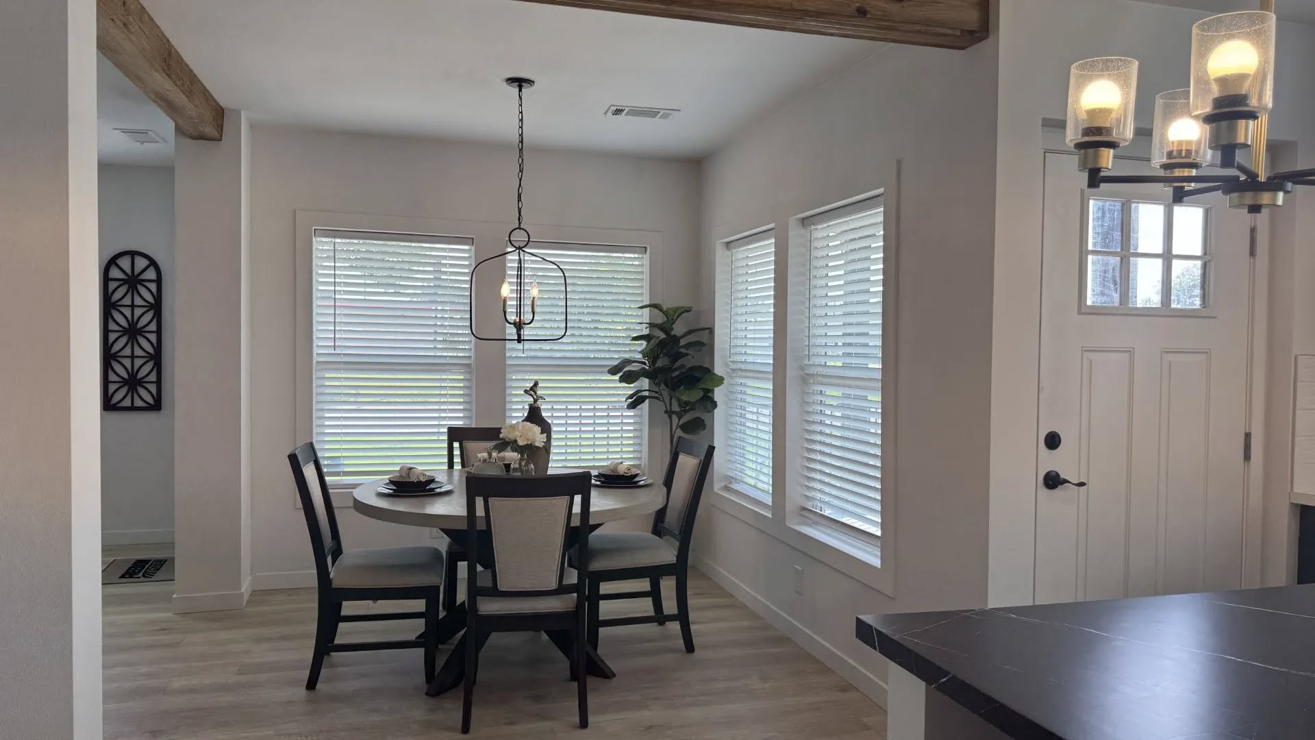 A cozy dining area with a round table set for four under a modern chandelier. Light streams in through several windows, creating a warm ambiance.