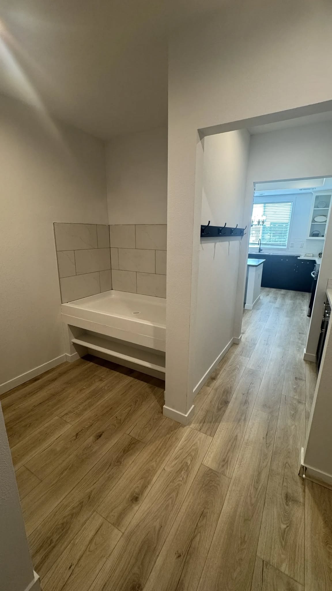 Narrow hallway with light wood flooring leads to a modern kitchen. A small nook with a tiled platform is on the left. The space feels clean and minimalistic.