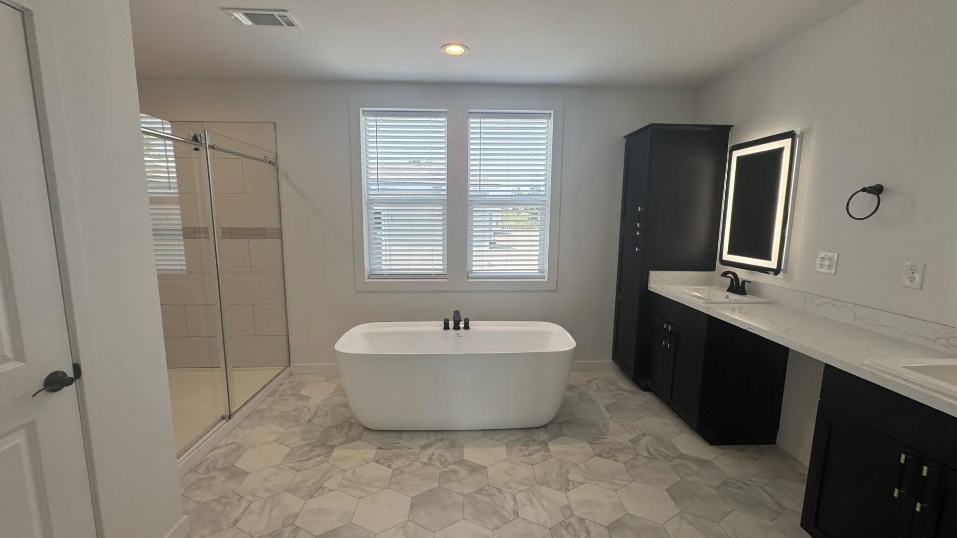 Spacious modern bathroom with a freestanding white tub, glass shower, and double vanity with a lit mirror. Hexagonal tile floor, neutral tones.