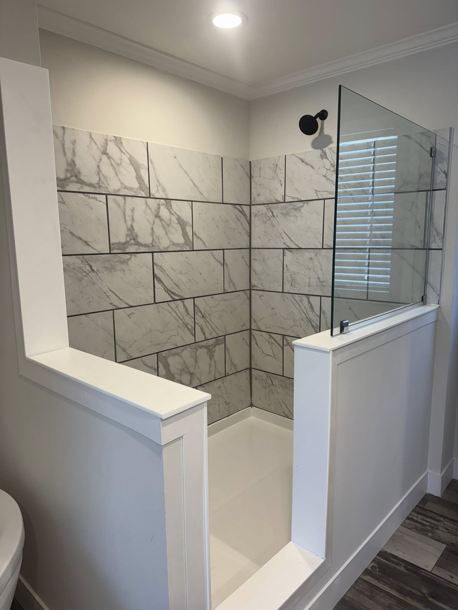 Modern shower with white marble tile walls and a partial glass partition. There's a recessed light above, creating a clean, minimalist feel.