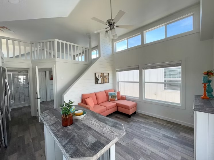 Bright, modern living space with a loft. Features a coral sofa, large windows, wooden floors, and a kitchen island; conveys a cozy, airy feel.