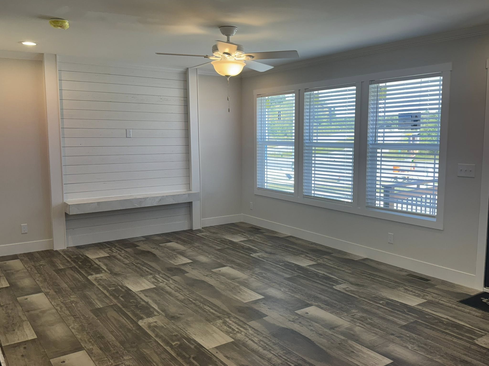 A bright, empty room with gray wood flooring and white walls. Large windows with blinds allow natural light. A ceiling fan with a light hangs overhead.