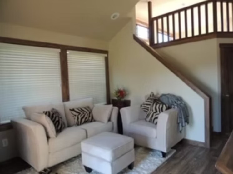 Cozy living area with beige sofas, zebra-patterned cushions, and an ottoman. Stairs with wooden railing lead to upper level. Bright and inviting atmosphere.