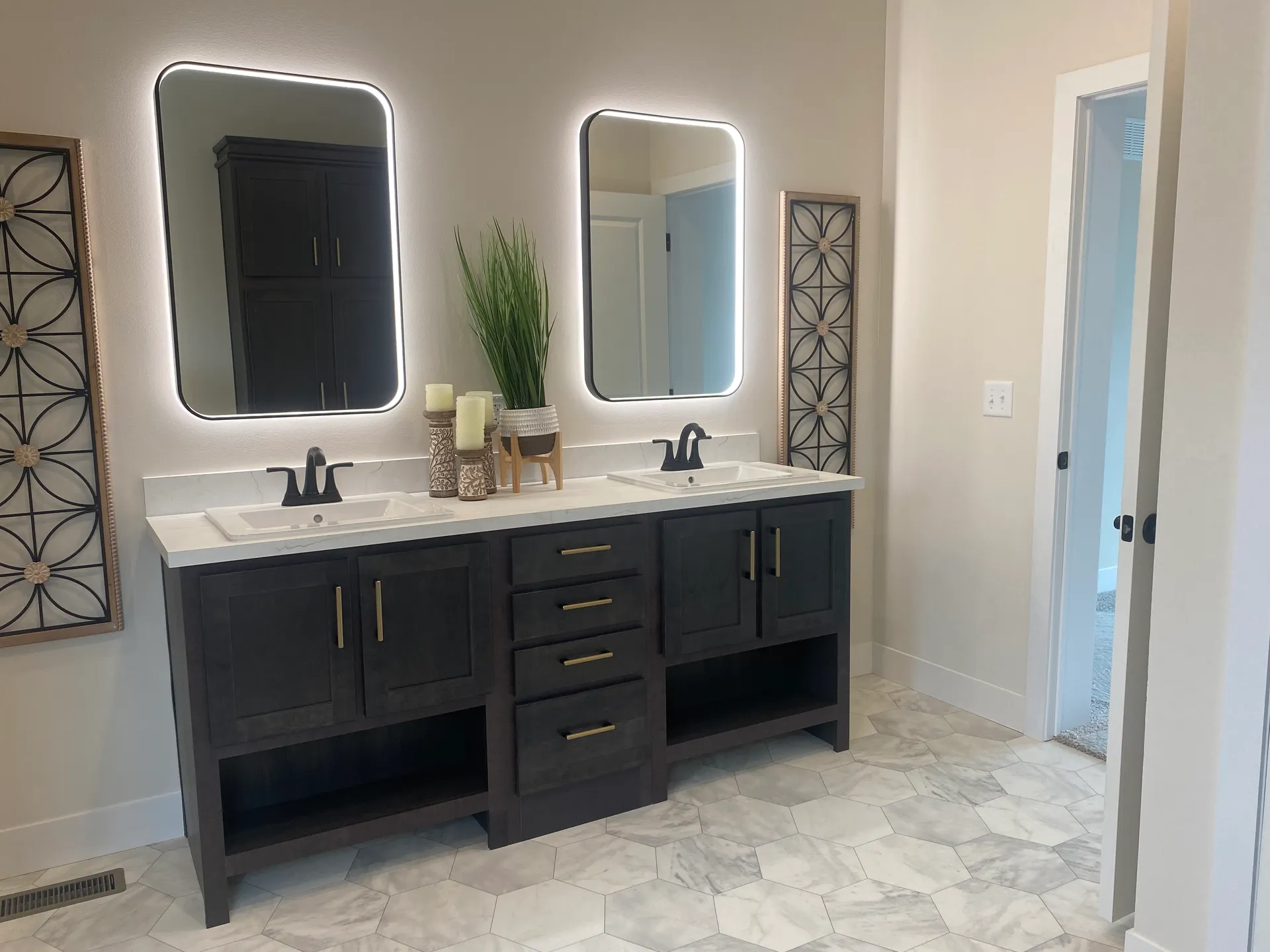 Modern bathroom with a dual-sink vanity featuring dark cabinets and gold handles. Two rectangular mirrors with backlighting are mounted above, flanked by decorative wall panels. A vibrant plant and candles adorn the countertop, while the hexagonal tiled floor adds elegance to the space.
