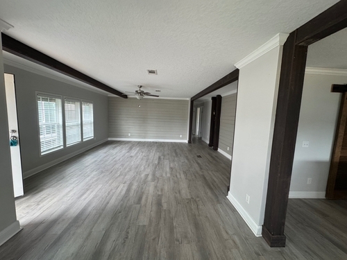Spacious empty living room with light gray walls, wood-style flooring, and large windows on the left. Dark wooden beams add contrast and elegance.