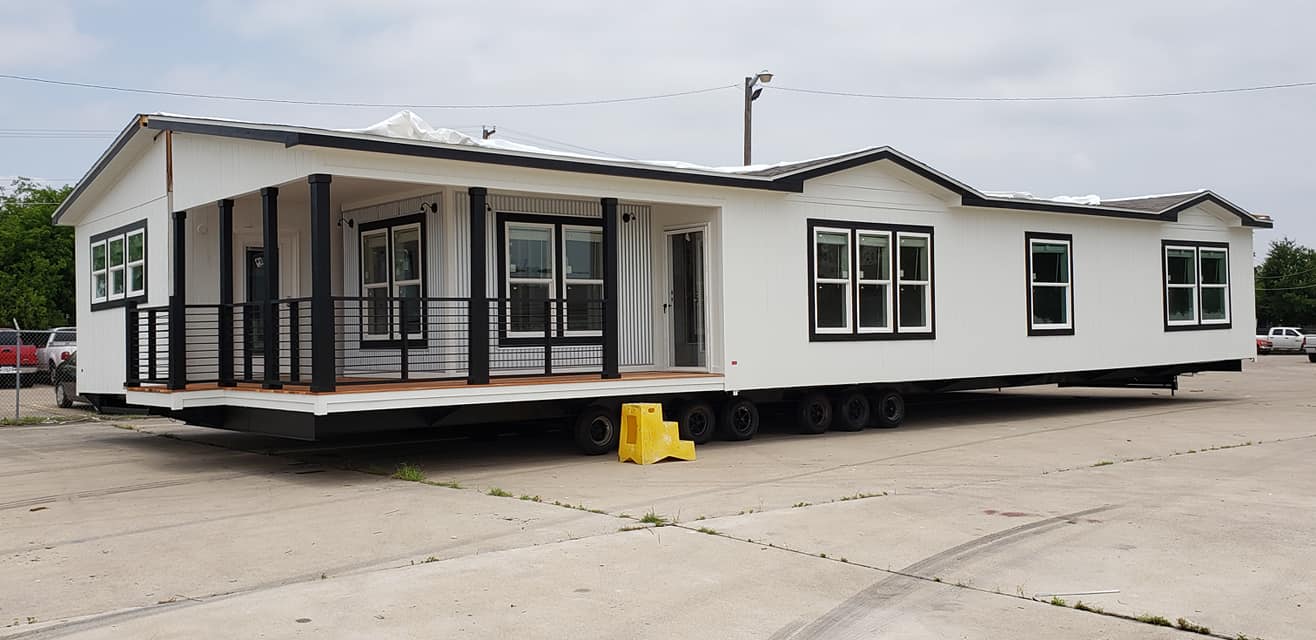 A large white mobile home on wheels with black-trimmed windows and a porch is parked on a concrete lot. The sky is cloudy, conveying a neutral tone.
