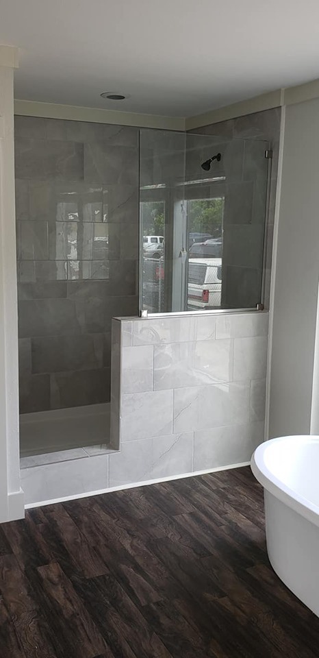 Modern bathroom with a glass-enclosed shower next to a freestanding white bathtub. The room has dark wood floors and light gray wall tiles.