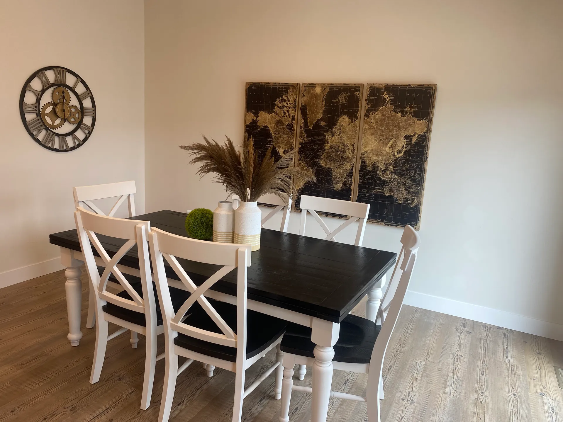 A dining room with a dark wooden table surrounded by white chairs, a wall clock, and a world map art piece. Decor includes a vase with pampas grass.