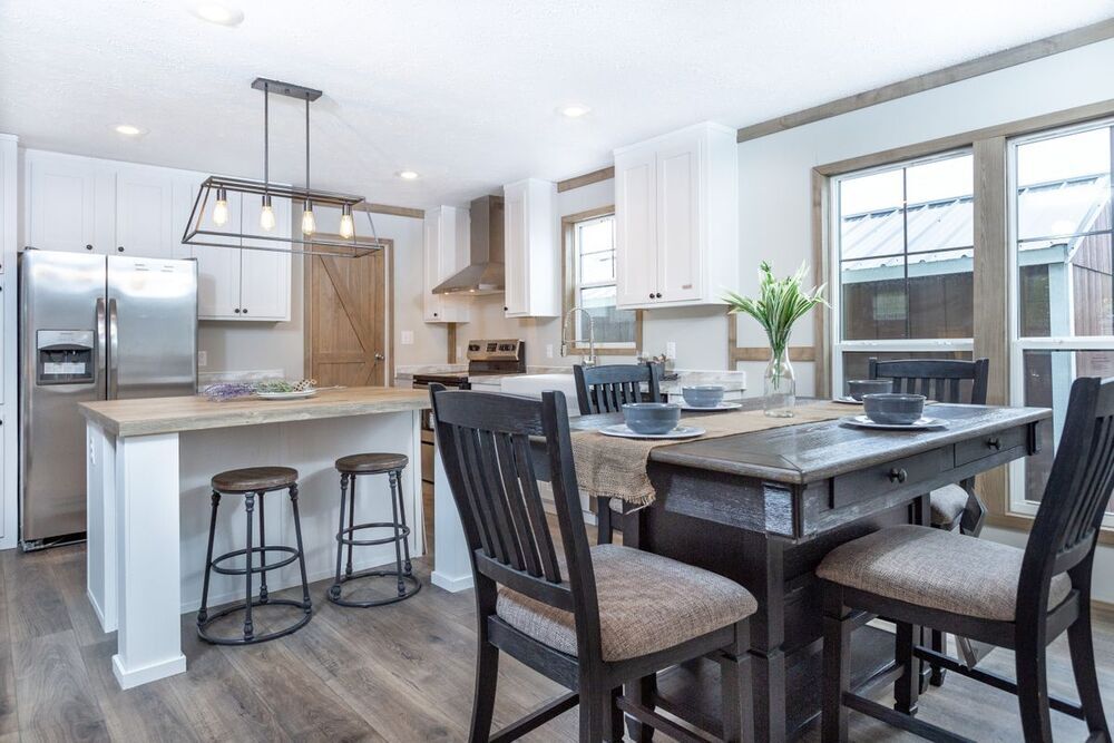 Modern kitchen with white cabinets, stainless steel appliances, and a wooden island with stools. A dark dining table with four chairs is set with plates. Bright, airy feel.