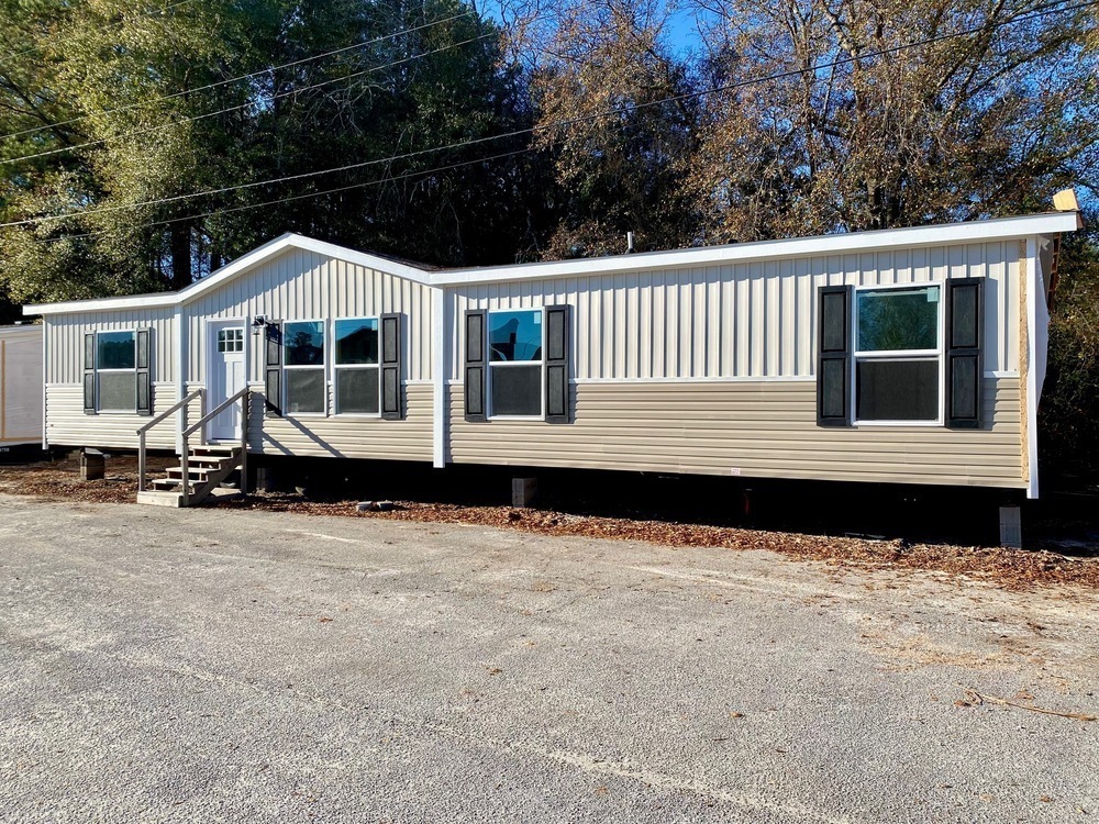 A beige, single-story mobile home with black shutters and multiple windows sits on a gravel lot. Leafless trees are visible in the background under a clear blue sky.