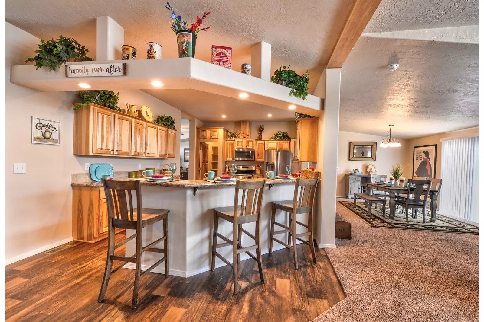 Warm, cozy kitchen and dining area with wooden cabinets and flooring. Bar stools line the counter. Dining table in background, soft lighting.