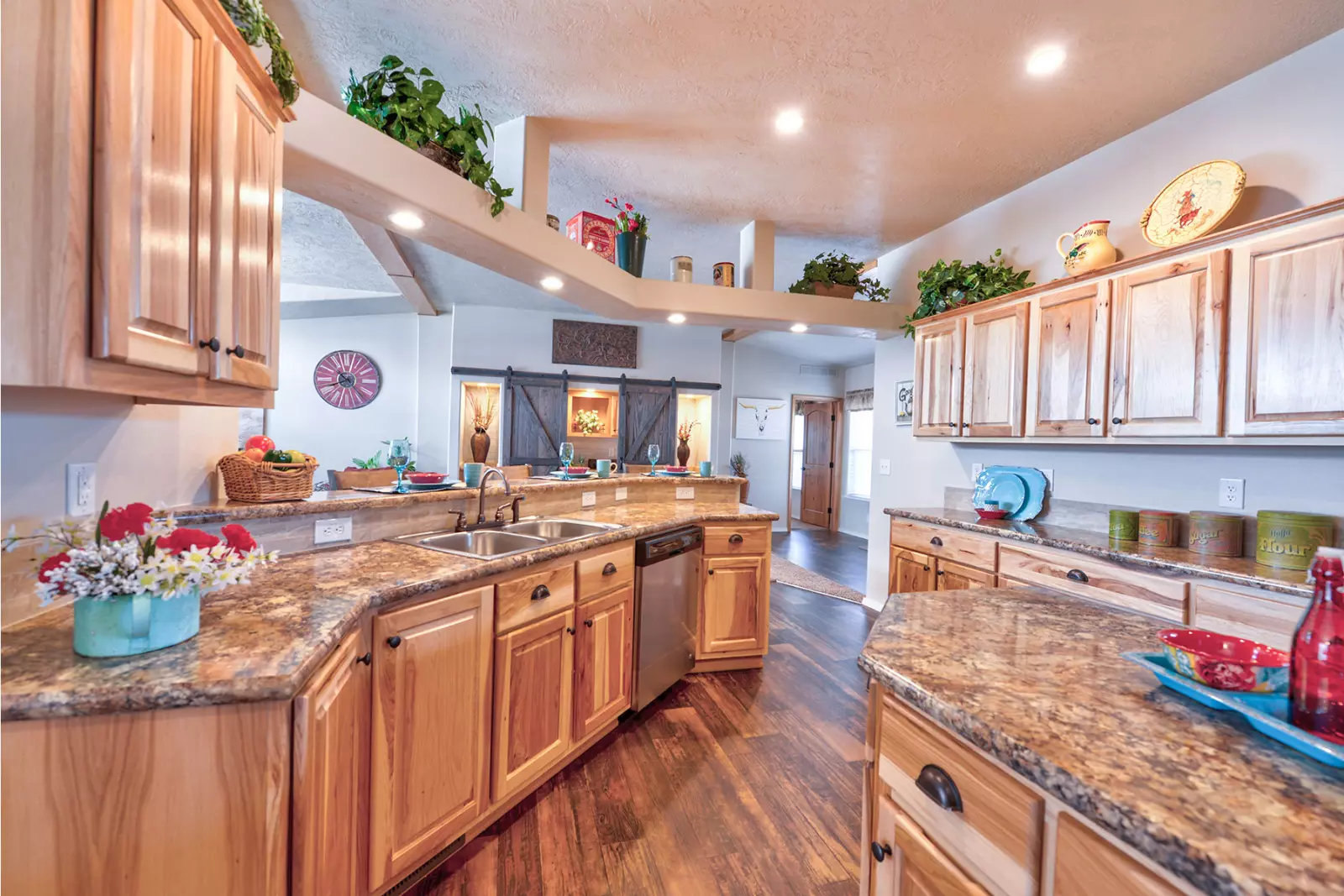 Spacious kitchen with wooden cabinets and granite countertops. Bright lighting highlights decorative plants and colorful dishware, creating a warm, inviting atmosphere.