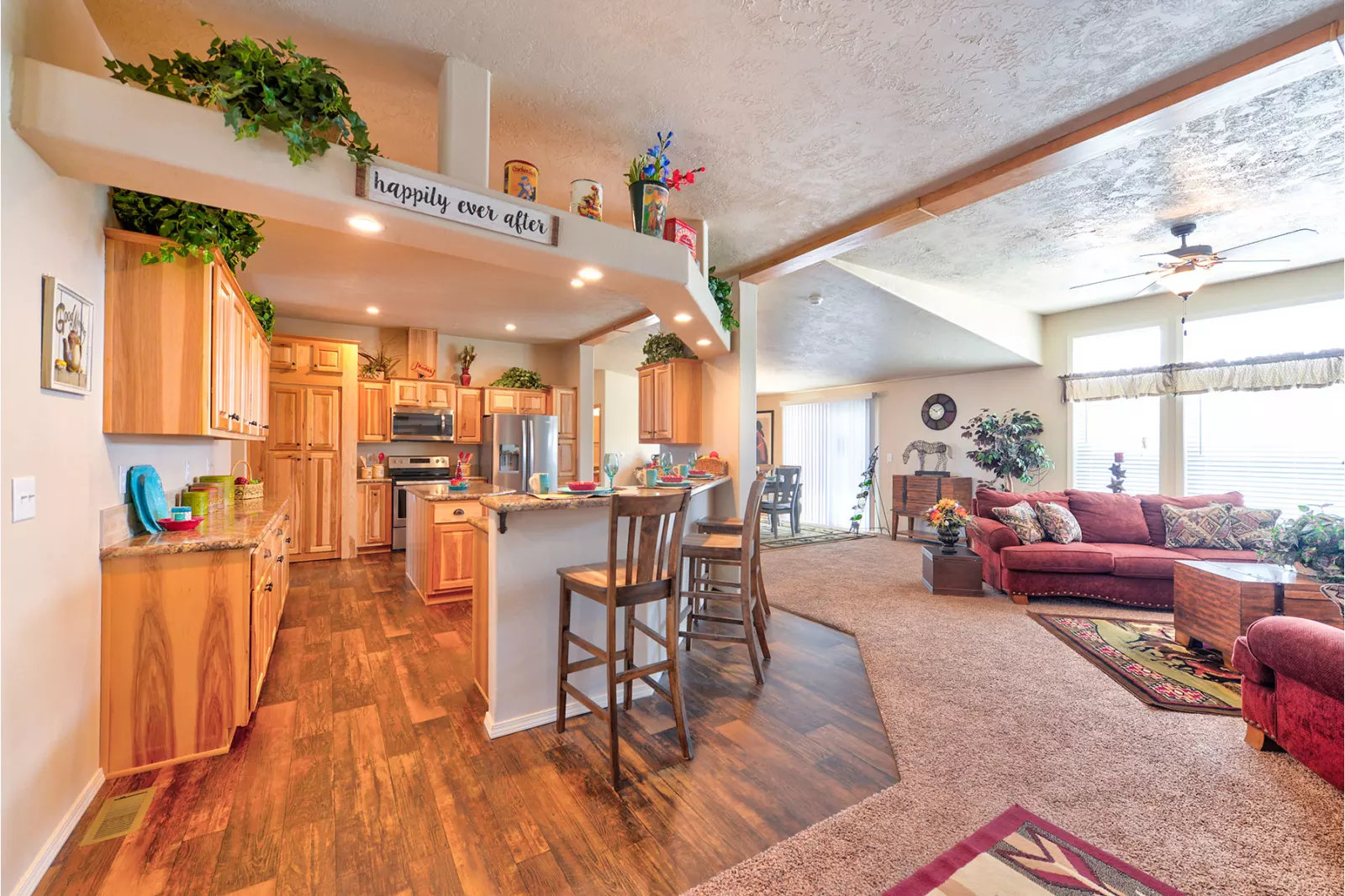 Bright, open-concept living and kitchen area with wood flooring. Features light wood cabinets, a bar with stools, cozy red sofas, and decorative plants.