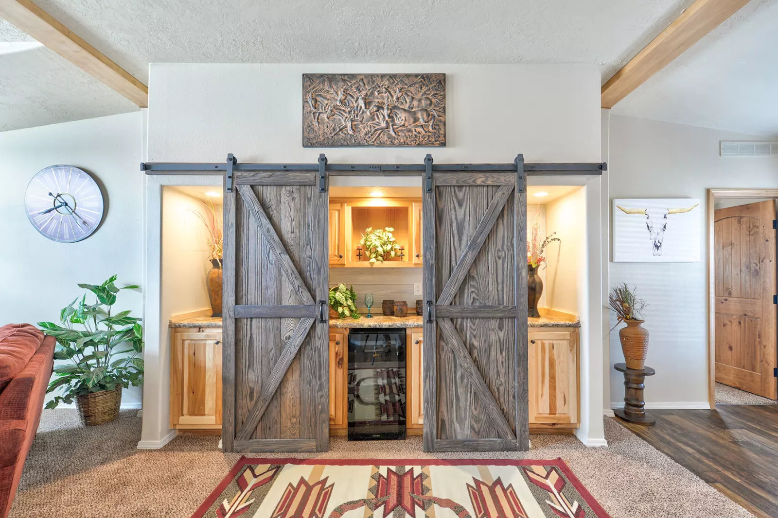 Rustic living room with sliding barn doors concealing a mini bar. Decor includes a wall clock, plant, horn art, and tribal rug, creating a cozy feel.