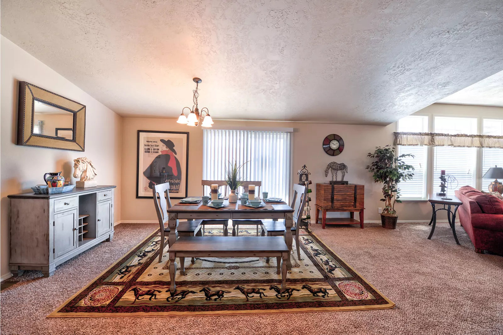 A cozy dining room with a wooden table and six chairs on a patterned rug. Soft lighting from a chandelier above, a sideboard with decor, and a red sofa in the background create a warm, inviting atmosphere.