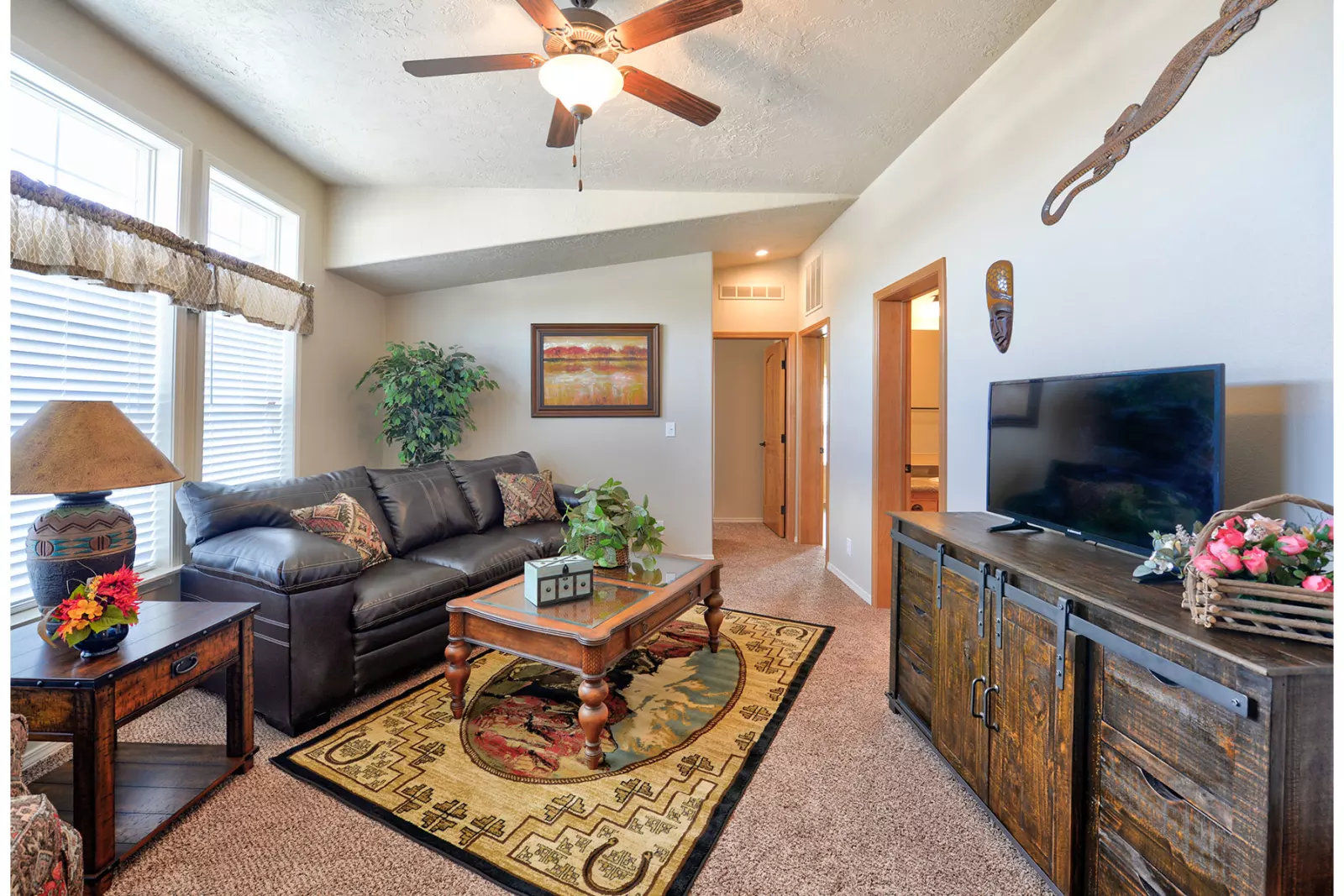 Cozy living room with a dark sofa, patterned rug, and wooden furniture. A ceiling fan, plants, and framed art add warmth. Natural light streams in.