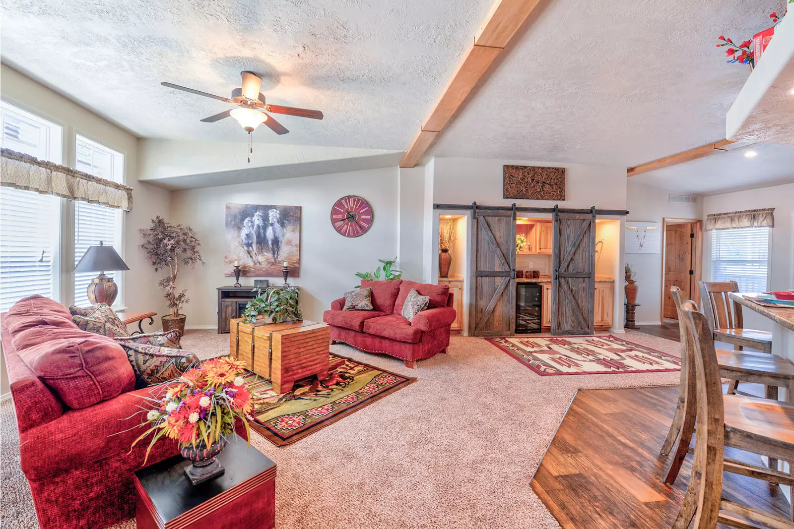 Cozy living room with red couches, rustic wood accents, and warm lighting. Features barn door cabinets, ceiling fan, and decorative plants. Inviting and homely atmosphere.