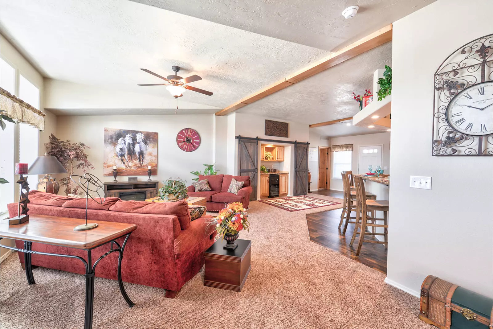 Spacious living room with red sofas, a ceiling fan, and warm lighting. Decor includes a wall clock, artwork, and plants, creating a cozy atmosphere.