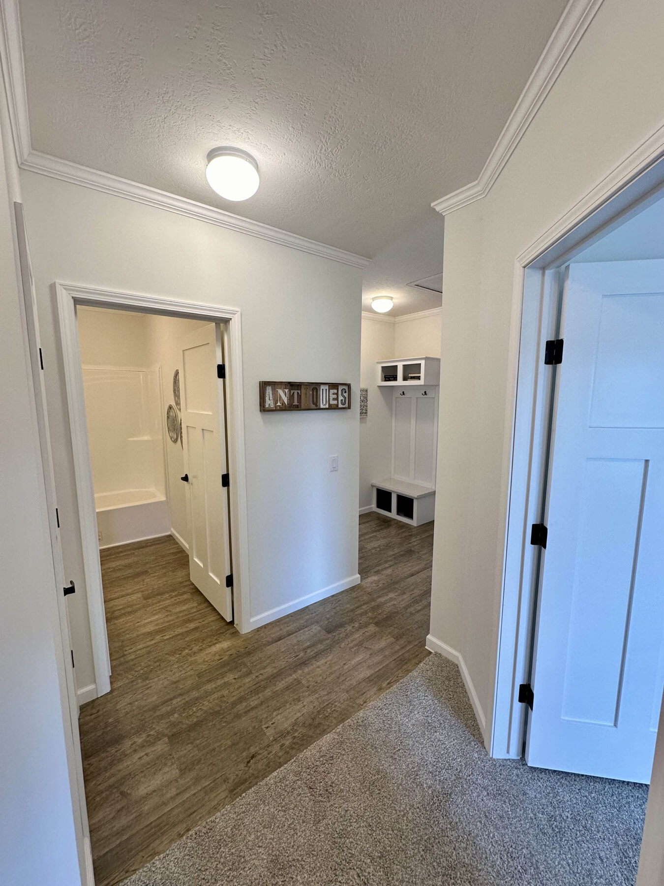 Hallway with wood and carpet flooring, leading to a bathroom and mudroom. Walls are white, featuring a sign reading "ANTIQUES," creating a cozy, welcoming tone.