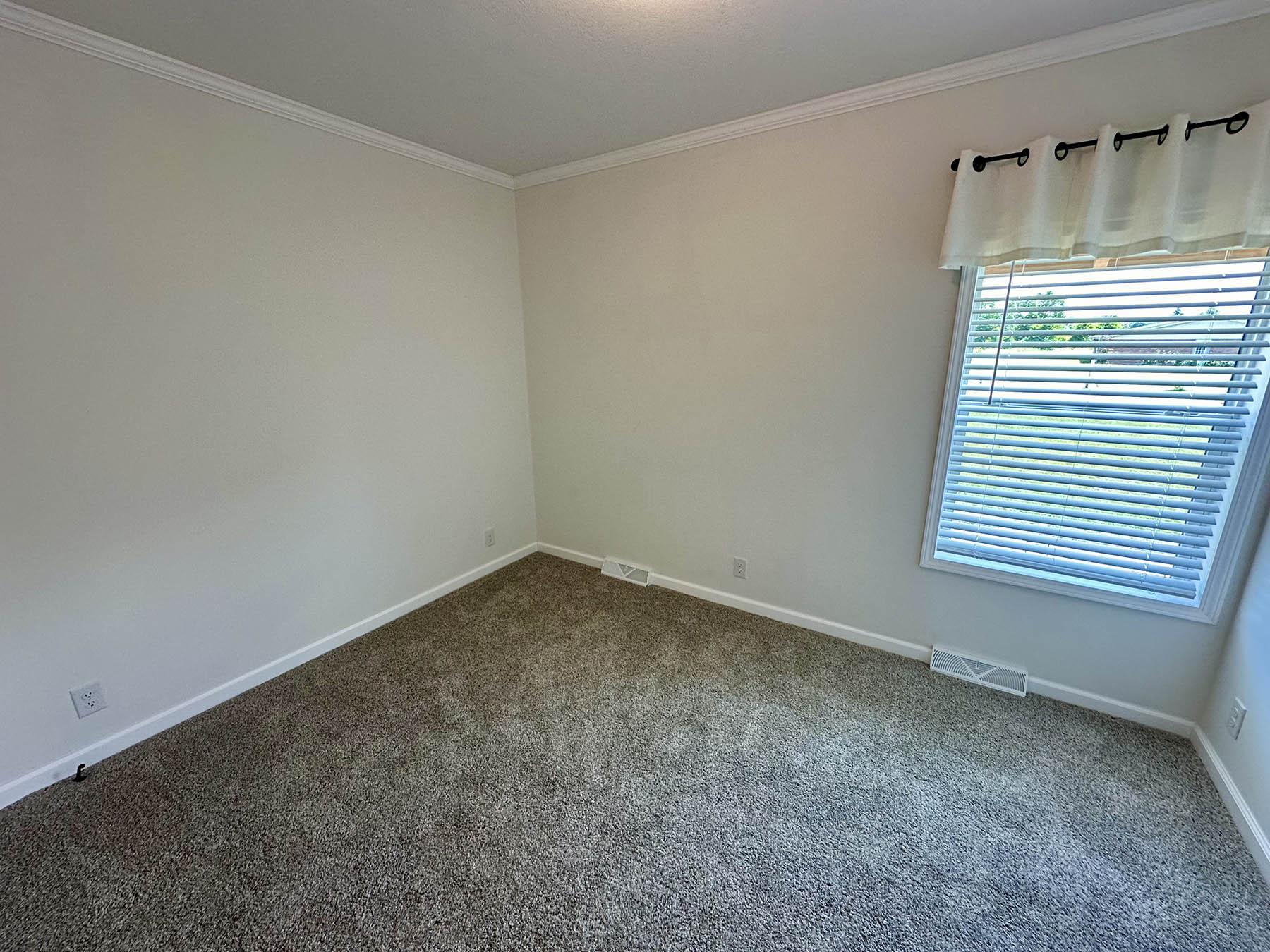 An empty room with beige carpet and off-white walls is shown. It has a large window with blinds and a white valance, letting in natural light. The room feels clean and minimalist.