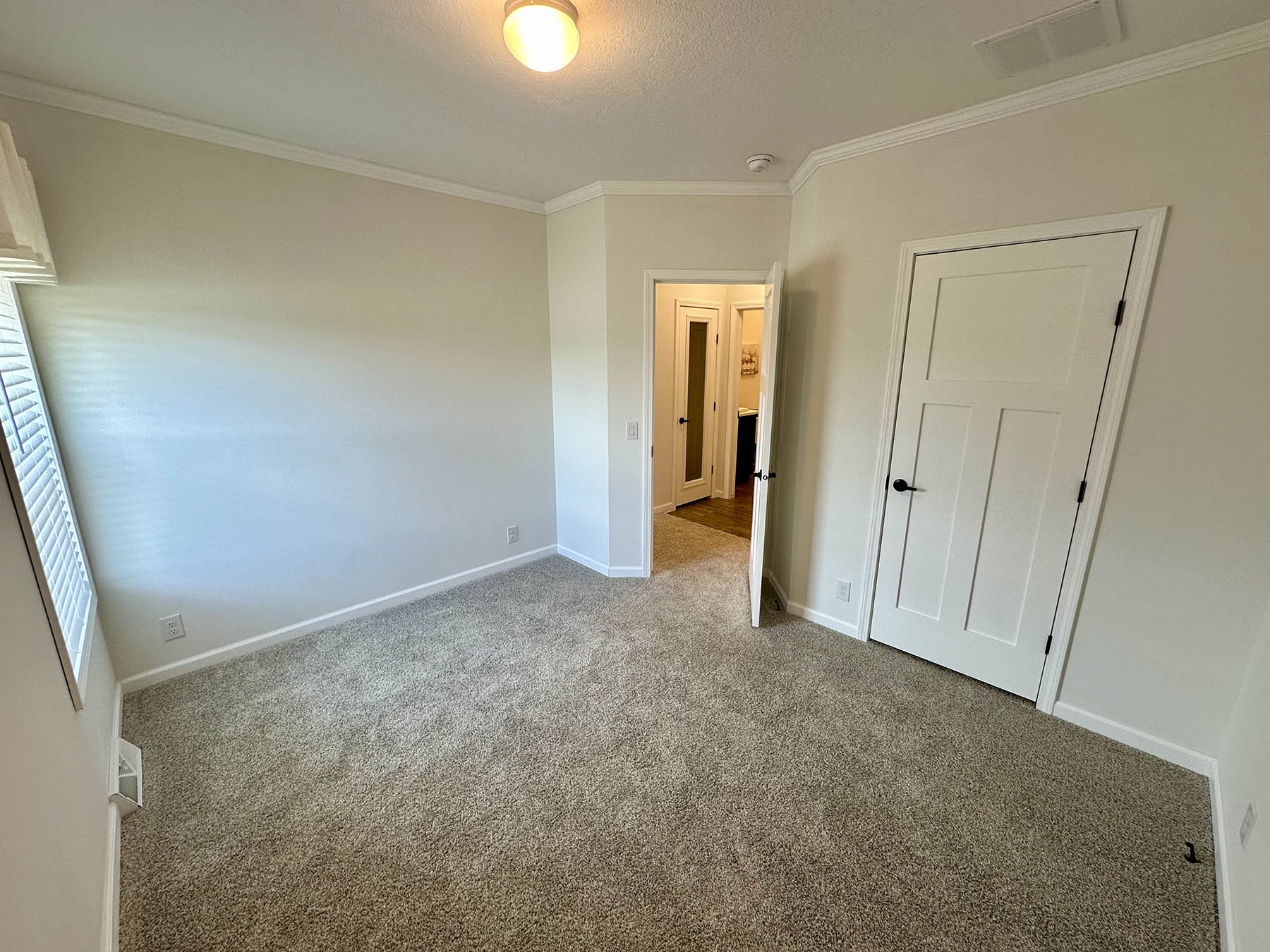 A small, empty room with beige carpet and light gray walls. There's a window on the left, a ceiling light, and two open white doors leading to a hallway.
