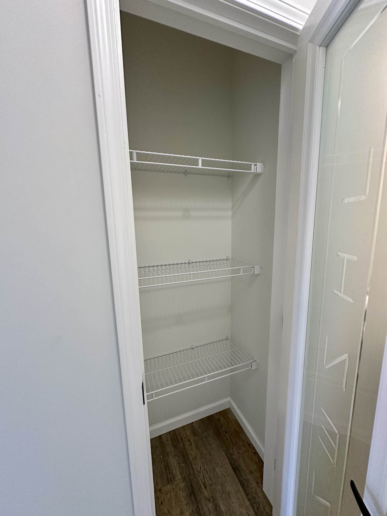 Empty closet with two white wire shelves, wooden floor, and white walls. The open door has glass with partial lettering visible, conveying a clean, minimalistic vibe.