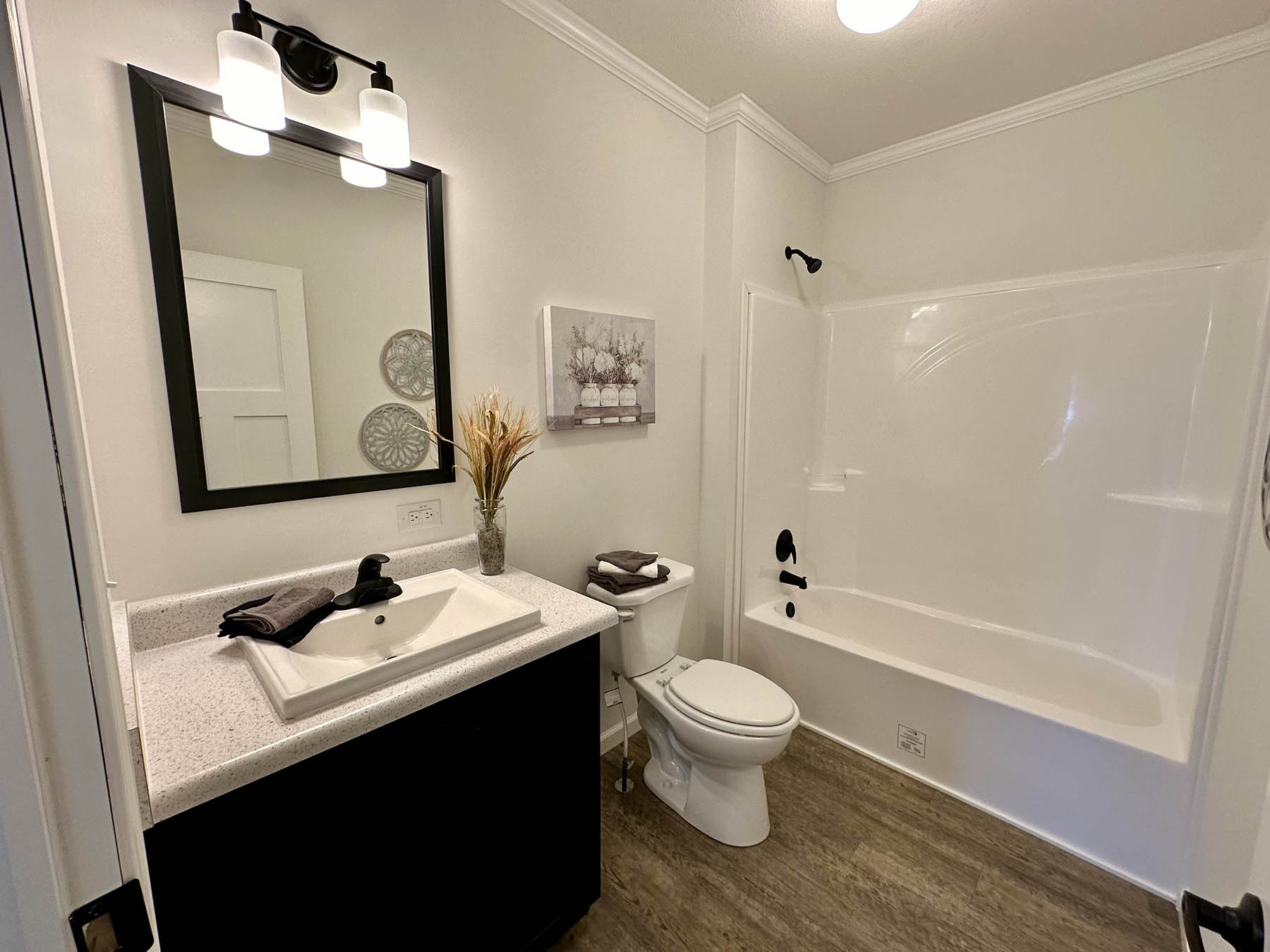 Modern bathroom with white walls, featuring a dark vanity and mirror, dual lights, toilet, bathtub, decorative art, and wooden floor. Clean and inviting.
