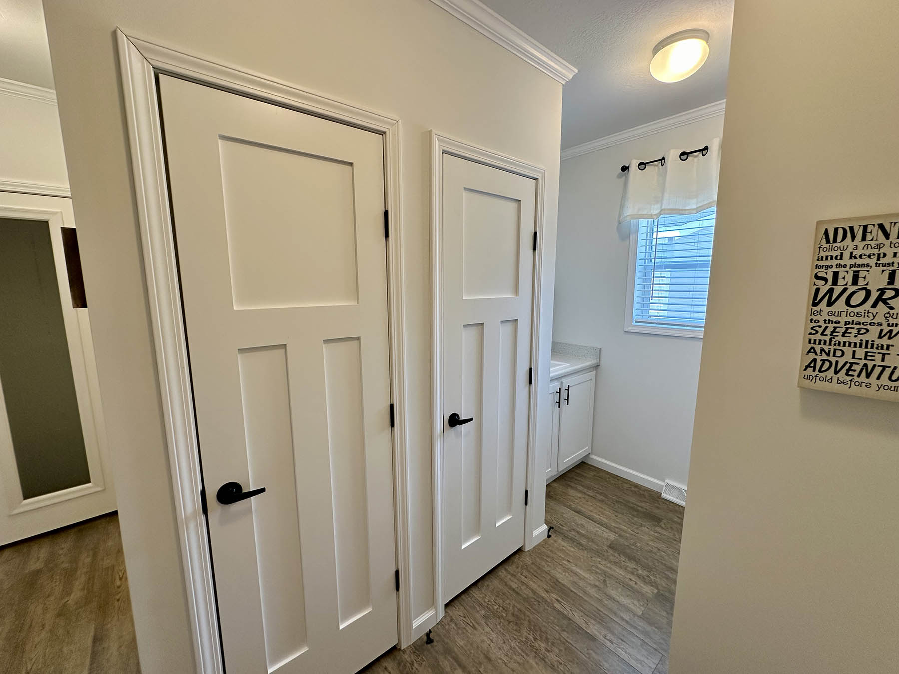A hallway with two white doors featuring black handles on the left. A small window with curtains illuminates the space. A framed motivational sign hangs on the right wall.