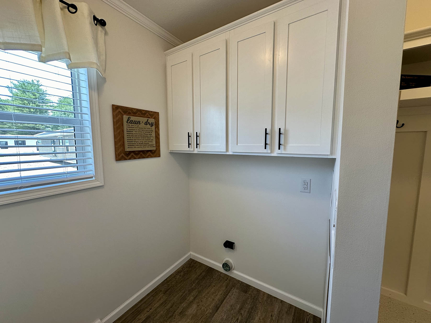 Laundry room with white cabinets and wood flooring. A sign reading "Laundry" is on the wall. A window with white curtains lets in natural light.