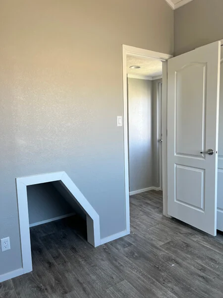 A small room with light gray walls and wood-patterned flooring. An open white door shows a hallway. A triangular alcove is visible on one wall.