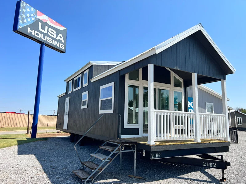 A modern tiny house with a dark exterior and large windows sits on a gravel lot under a clear blue sky. A "USA Housing" sign stands nearby.