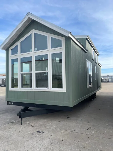 A green modular house with large, white-framed windows sits on a paved lot under a blue sky. The house has a gabled roof and a modern design.
