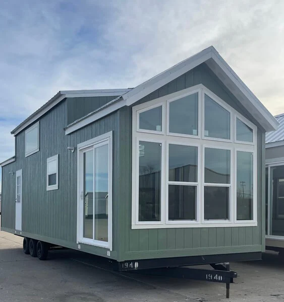 Green tiny house on wheels with large front windows, set against a cloudy sky. The modern design and clean lines evoke a minimalist, cozy feel.