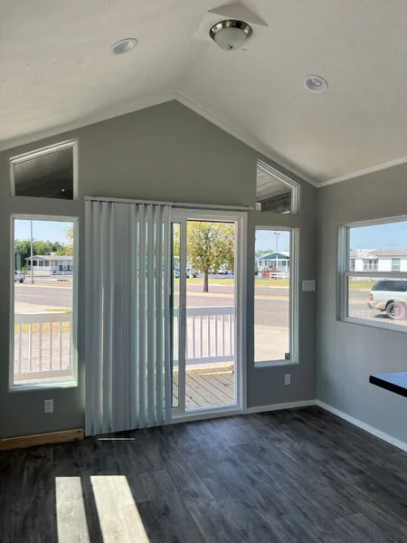 Inside a sunlit room with gray walls and dark wood floors, large windows and glass doors open to a porch with a bright, peaceful street visible outside.