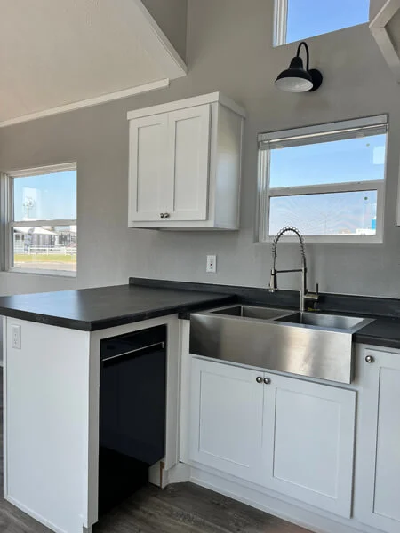 Modern kitchen with white cabinets, stainless steel farmhouse sink, and black countertops. Bright light from windows illuminates the space.