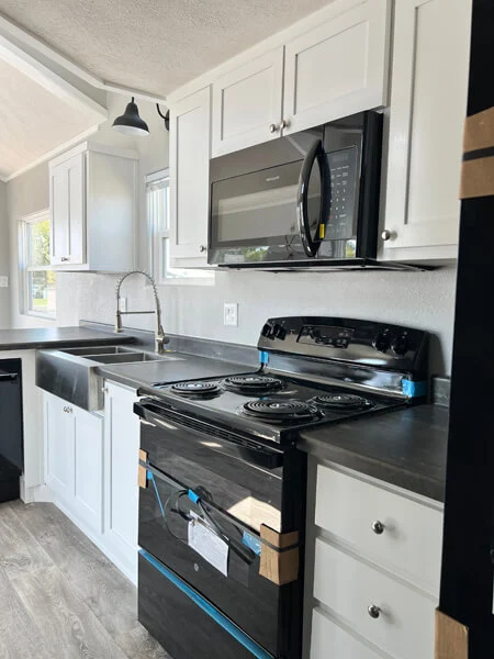 Modern kitchen featuring white cabinets, a sleek black stove with protective film, and a microwave. Sunlit space with a minimalist, clean feel.
