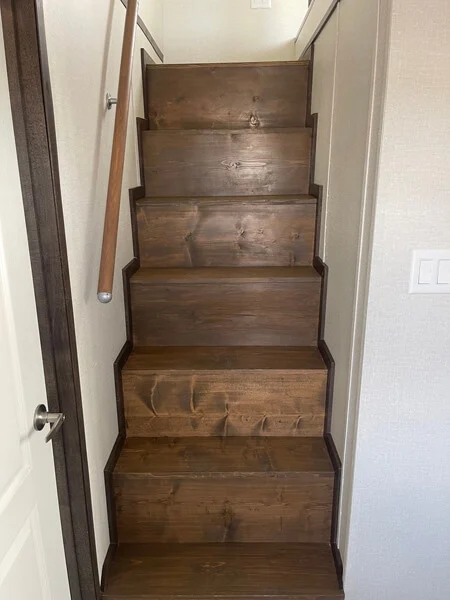Narrow wooden staircase with six steps leading up, flanked by white walls. A simple handrail runs along the left side, emitting a cozy, rustic vibe.
