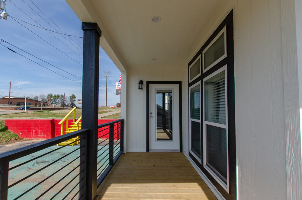Covered porch with wooden flooring and railing, leading to a glass door. Large windows on the right, with a red wall and blue sky outside.