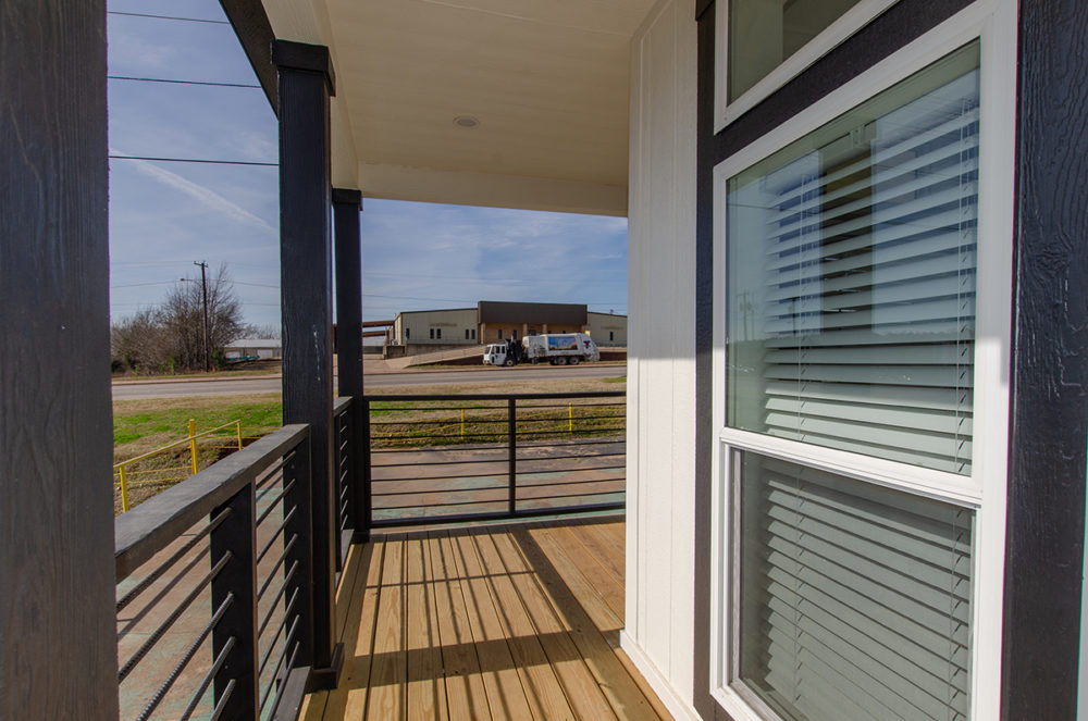 A wooden porch with black railings overlooks an industrial area under a blue sky. A parked truck is visible in the distance, creating a calm atmosphere.