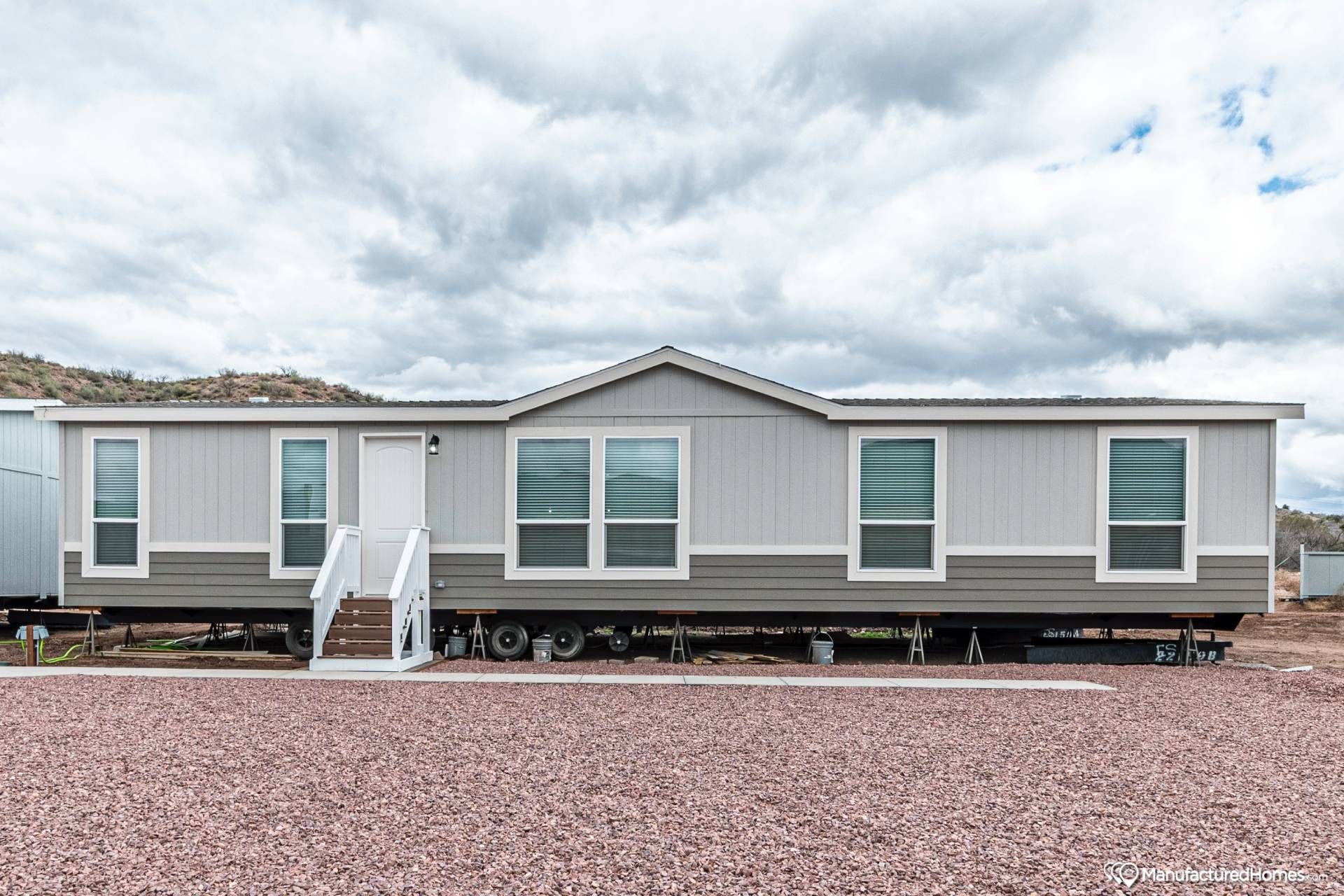 A modern manufactured home with gray siding and white trim sits elevated on wheels. It features several large windows and a small staircase at the entry. A cloudy sky creates a neutral backdrop.