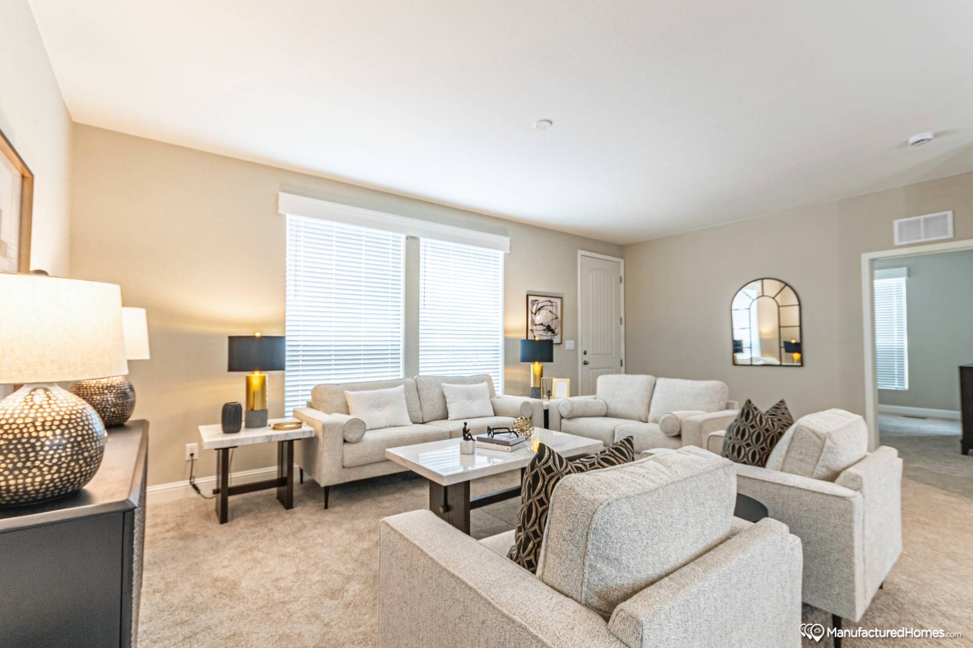 Modern living room with beige sofas, a coffee table, and decorative pillows. Soft lighting from lamps and a large window create a cozy atmosphere. A mirror and framed art adorn the walls.