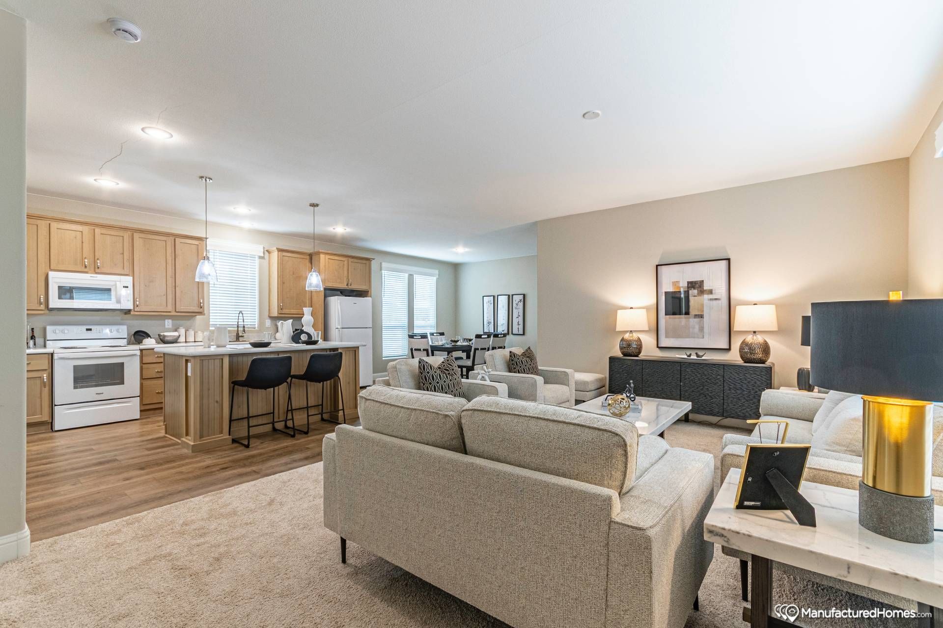 Spacious open-plan living room and kitchen with beige sofas and modern decor. Wooden kitchen cabinetry, a dining area, and warm ambient lighting.