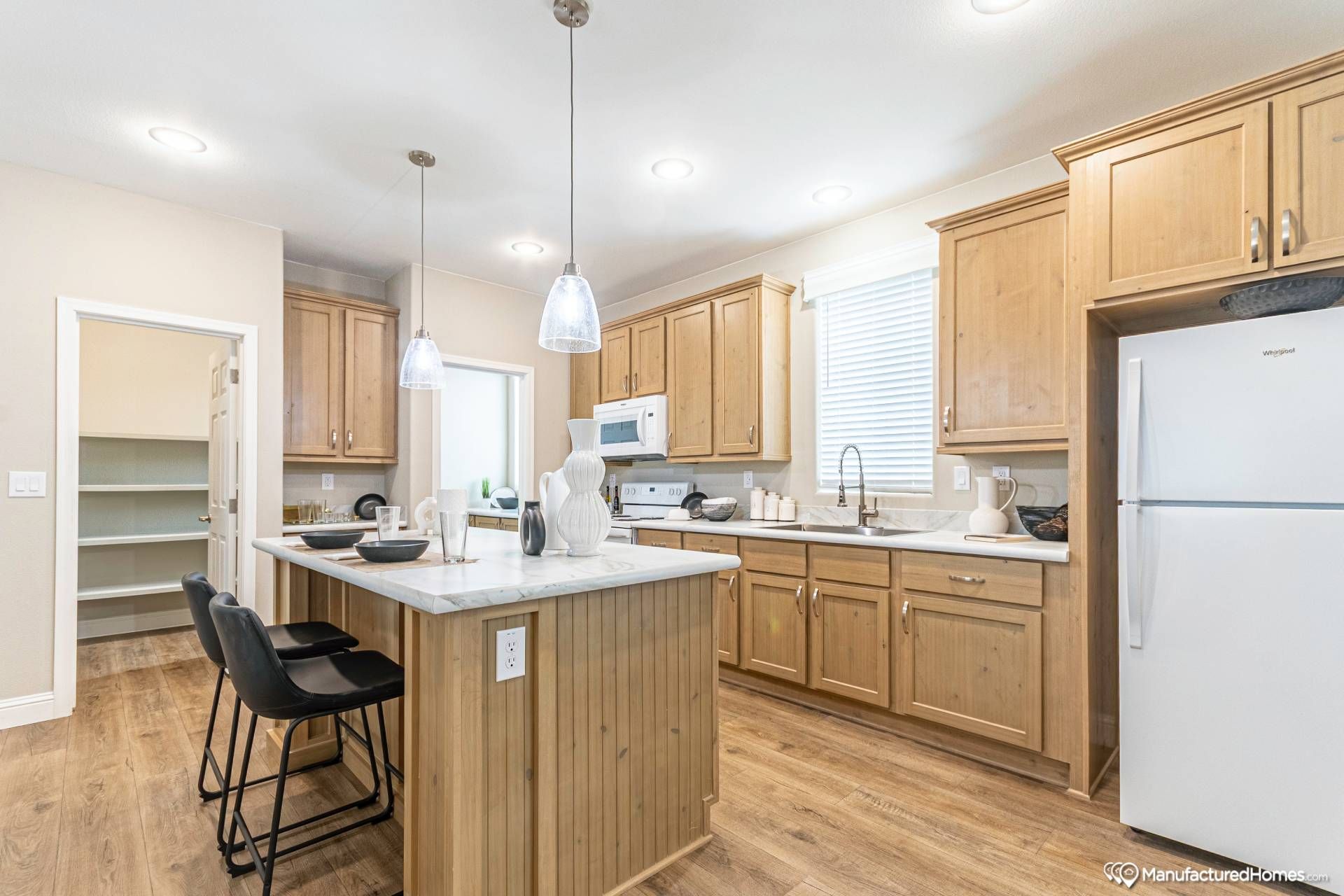 Bright kitchen with wooden cabinets, a marble island, and two black stools. Pendant lights hang above. Refrigerator and microwave are visible. Warm and inviting atmosphere.