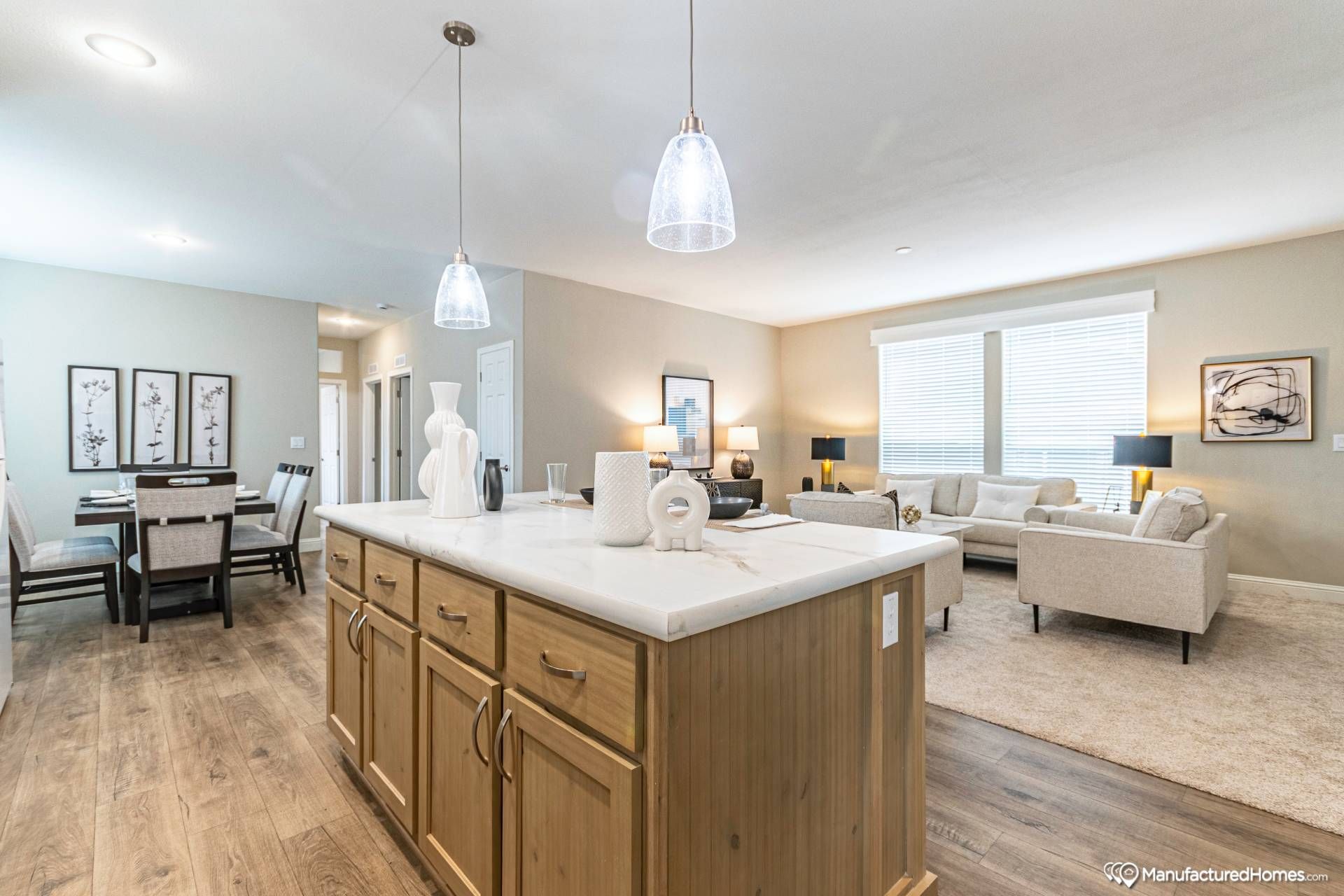 Bright, modern open-concept living area with wooden floors, featuring a kitchen island with pendant lights, and a cozy living room with beige sofas.