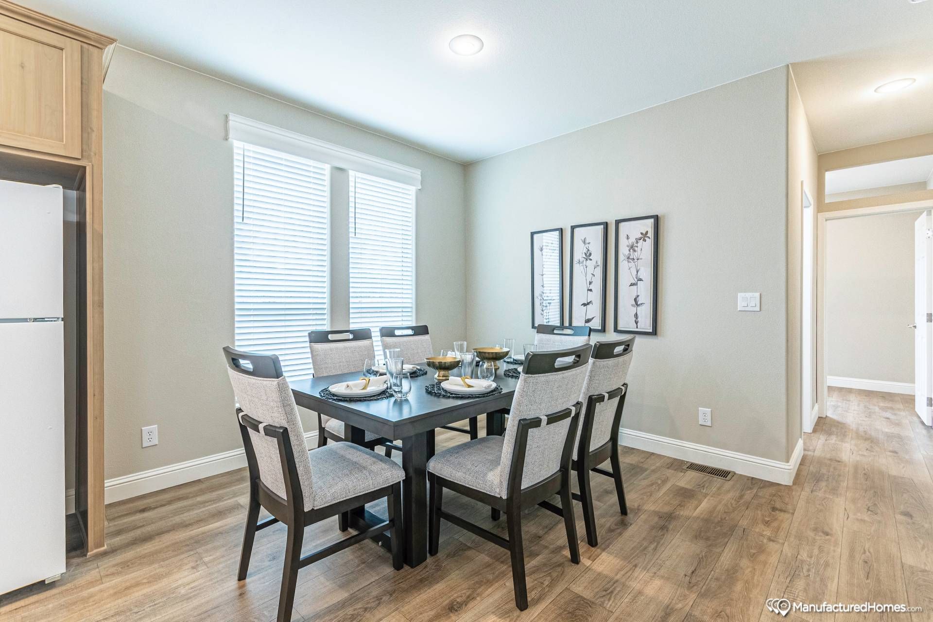 Modern dining room with a dark wooden table set for six. The room features light gray walls, wooden flooring, and three framed art pieces. Soft, inviting atmosphere.