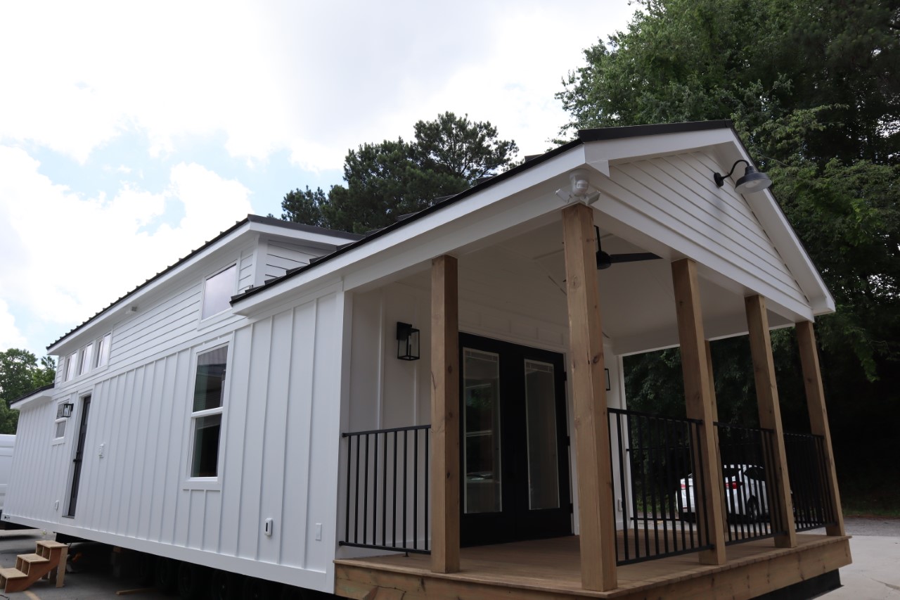 A modern tiny house with white siding, a small porch with wooden posts and black railings. The setting is outdoors with trees in the background.