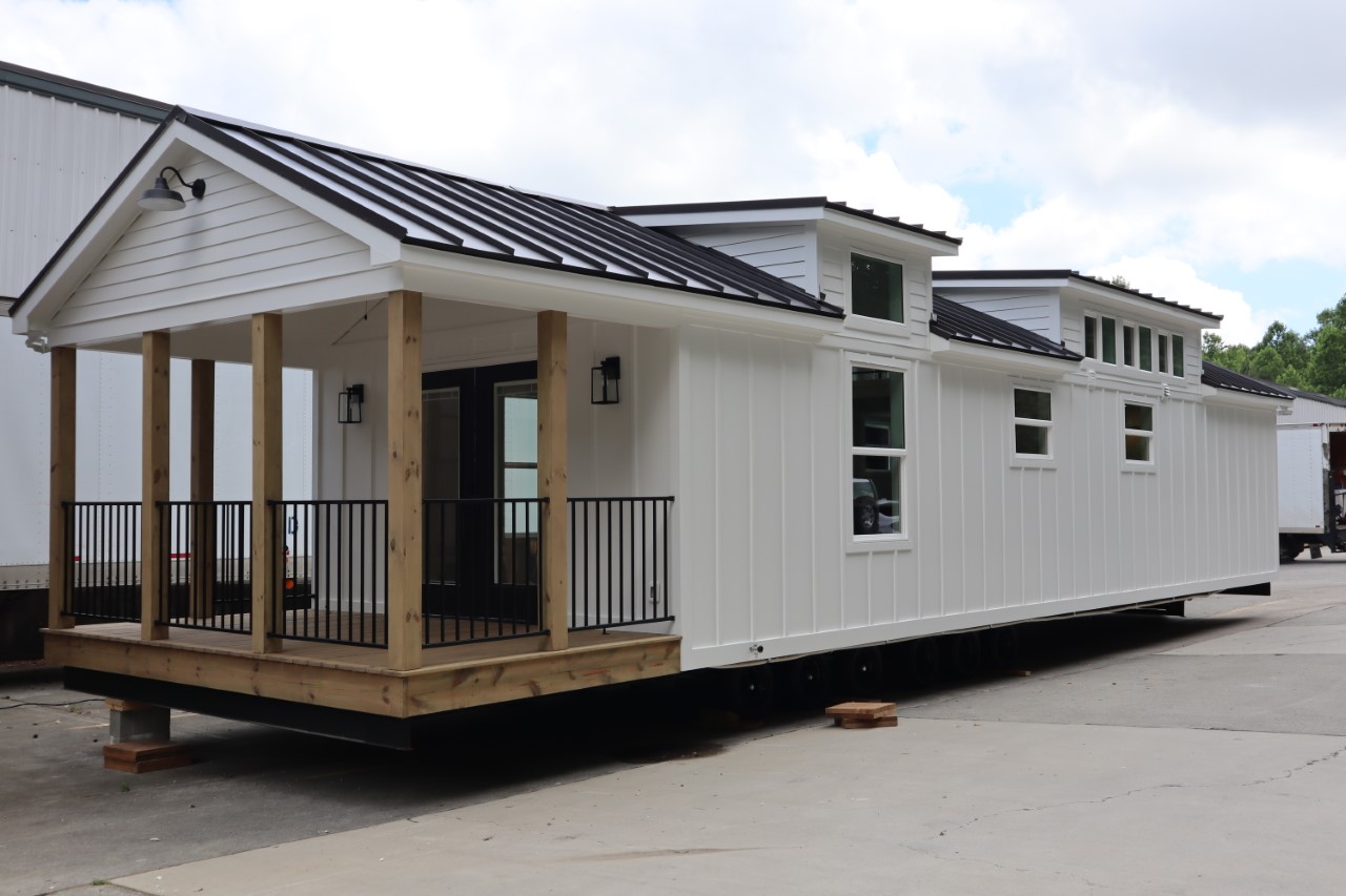 A white tiny house with a wooden porch and black metal roof sits on a concrete lot. It features multiple windows and a welcoming, modern design.