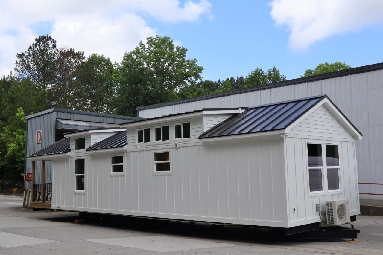 A modern white tiny house with a metal roof is parked outdoors near a large industrial building. The scene is surrounded by trees and a blue sky.