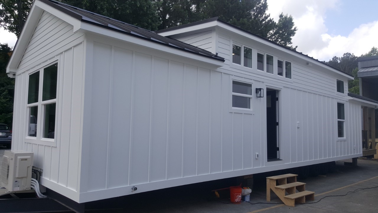 A modern white tiny house with a slanted roof sits on a lot. Large windows and solar panels suggest energy efficiency. Wooden steps lead to the entrance.