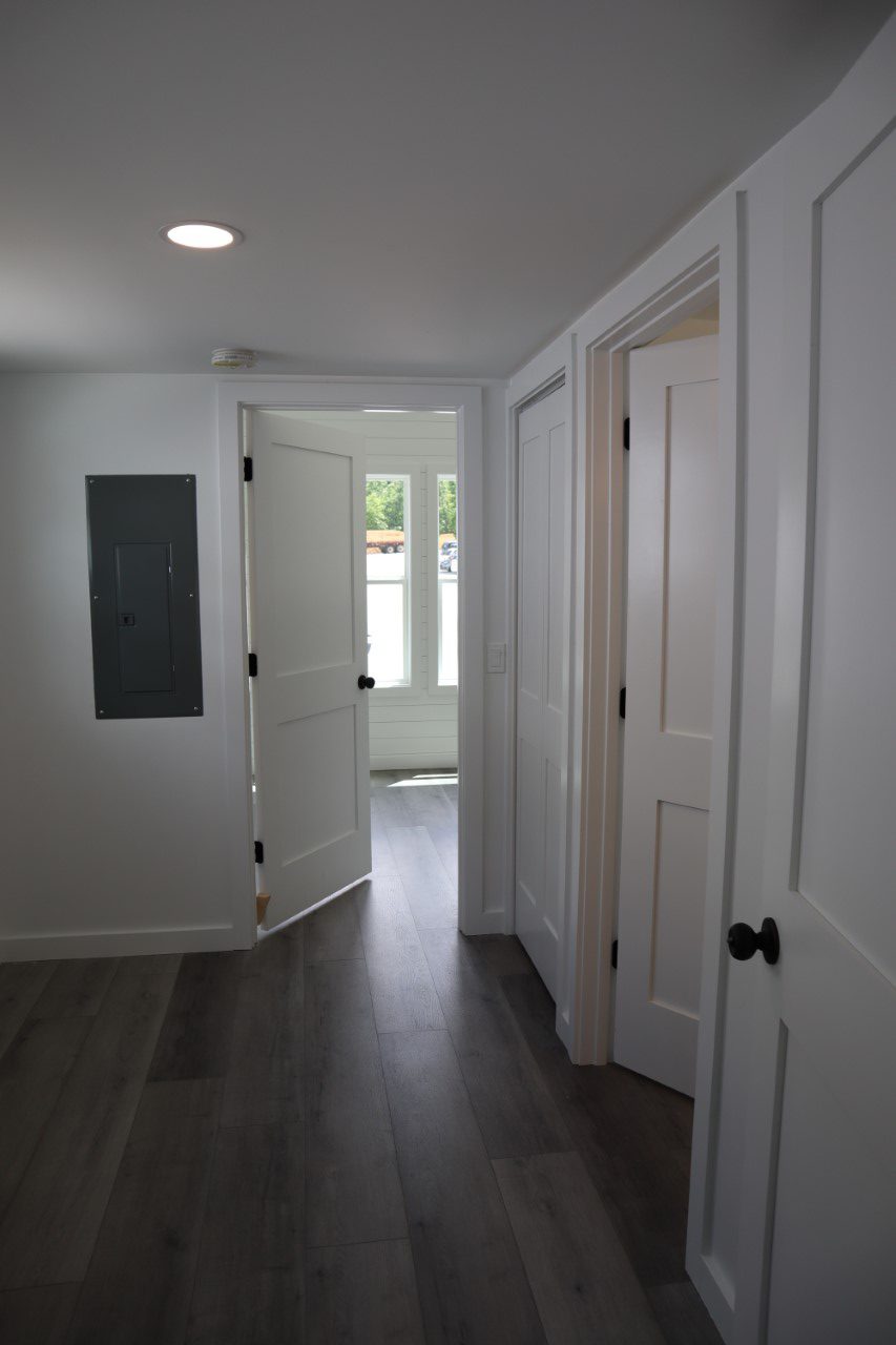 Hallway with dark wood flooring and white walls. Four white doors are visible, two ajar, leading to a sunlit room with large windows. Calm and inviting.